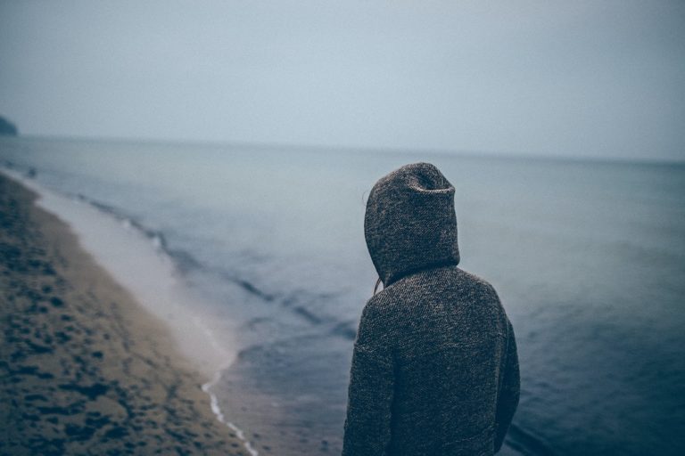 man with hoodie on with his back facing the camera overlooking the beach on a gloomy day to symbolize depression and addiction