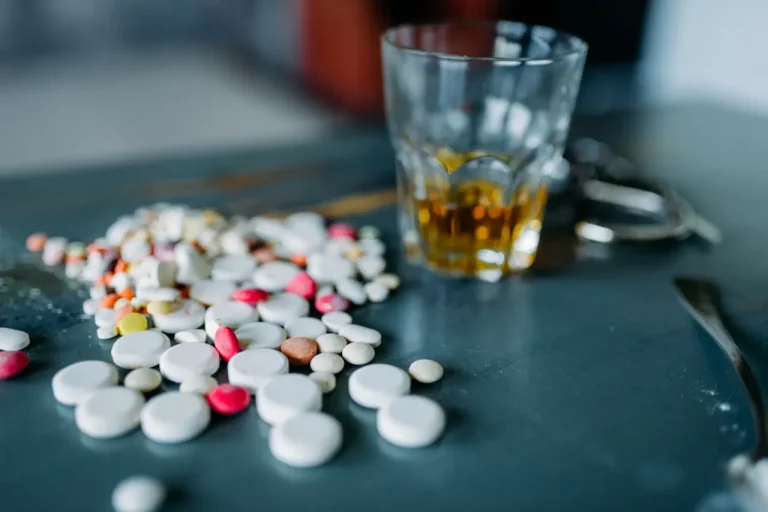 A glass of alcohol on the table with a lot of medication pills scattered on the table.